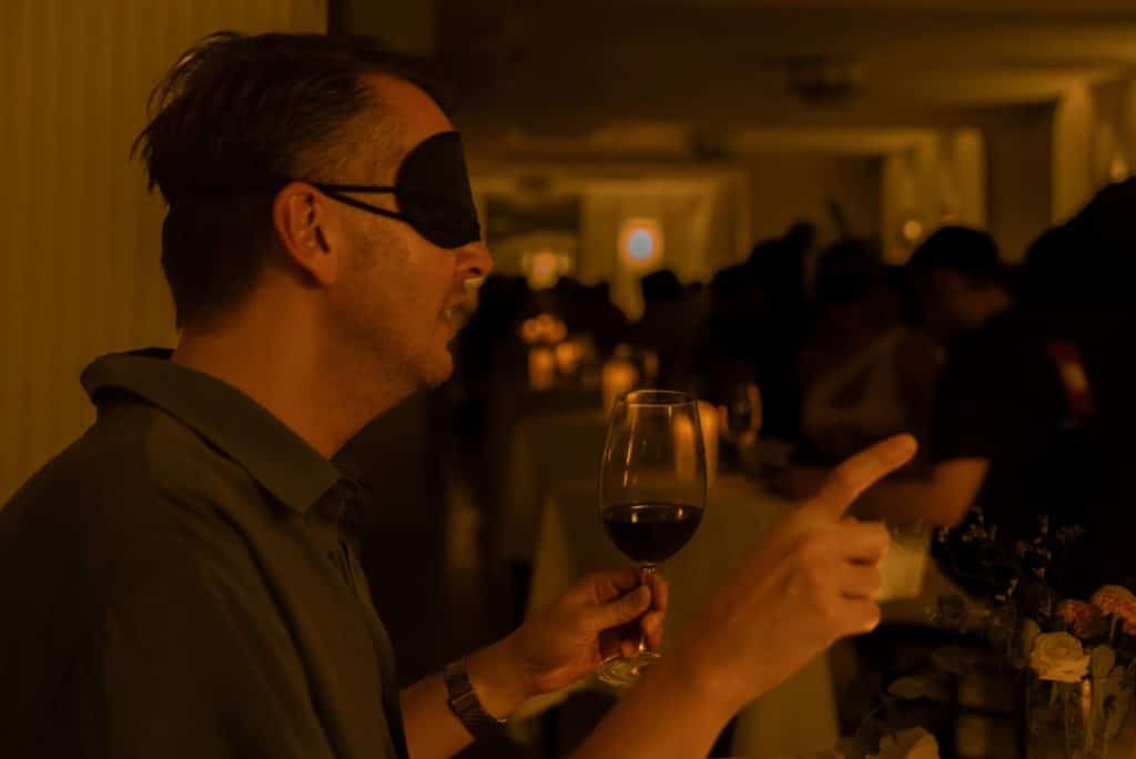 A man with a blindfold on holding a glass of wine and pointing his finger