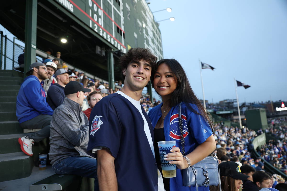A lively crowd of Chicago Cubs fans fills the iconic Wrigley Field bleachers on a sunny game day, decked out in jerseys, caps, and team spirit. Fans cheer, raise their drinks, and soak up the energy of the ballpark. In the foreground, a couple pauses to snap a photo with the sea of fans behind them, capturing the perfect summer moment at Wrigley.