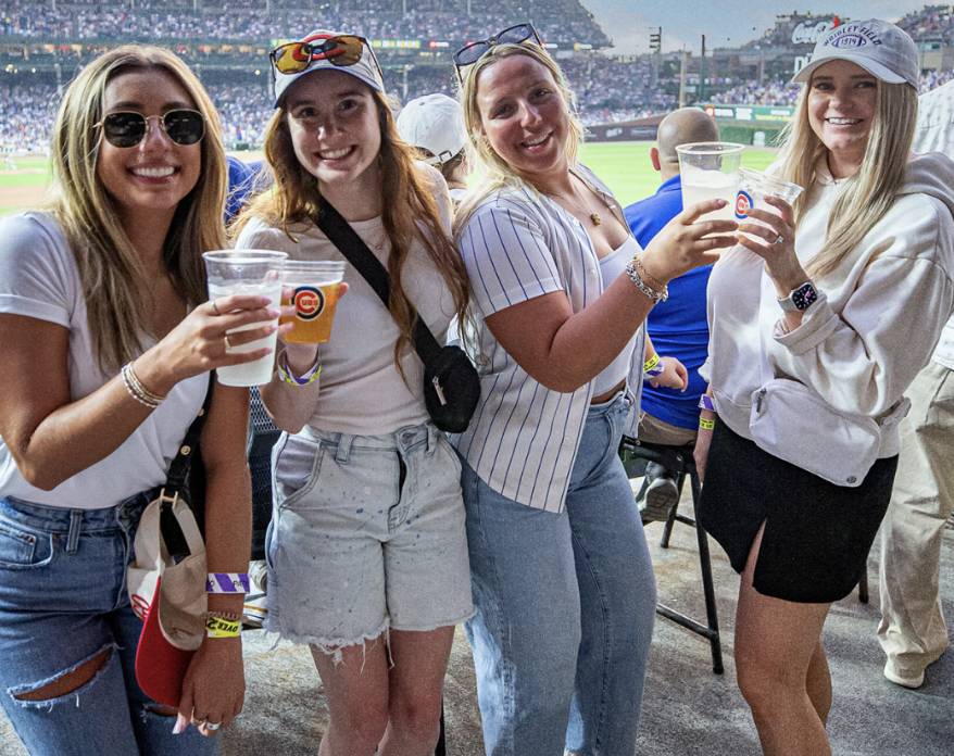 Four friends smile and pose with their free drinks at a sunny Chicago Cubs game, showing off the fun perk that comes with their ticket, including beer, wine, or a NÜTRL vodka seltzer.