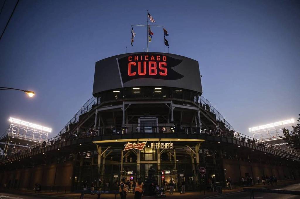 Al anochecer en el Wrigley Field, las luces del estadio brillan contra el cielo cada vez más oscuro, proyectando un tono dorado sobre el campo. Los aficionados se preparan para la velada mientras el emblemático marcador se ilumina y el perfil de la ciudad comienza a parpadear en la distancia.