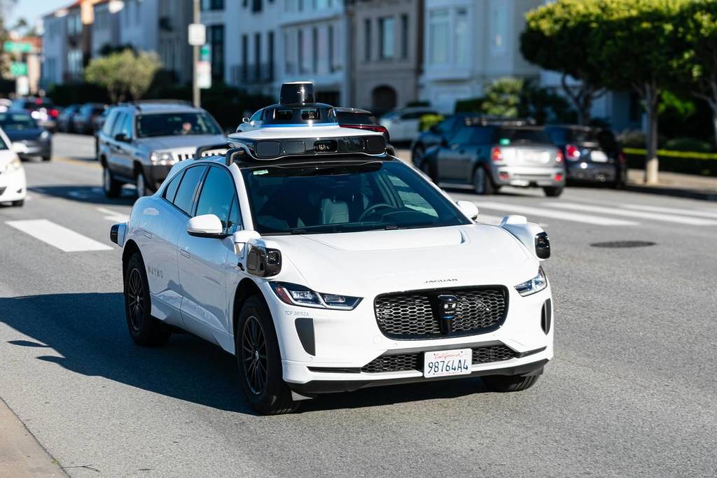 Waymo One Jaguar driverless autonomous car equipped with lidar sensors and cameras drives on a multi-lane city road.