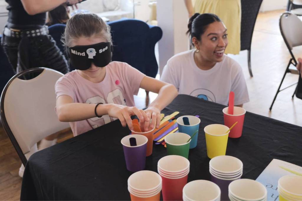 A child wearing a blindfold while picking up a cup as a woman smiles at Superminds Academy