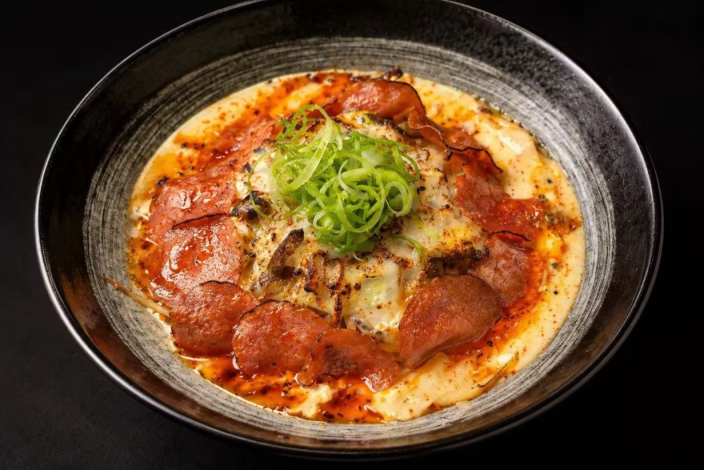 Strings Ramen's Deep Dish Ramen in a black bowl, with crispy pepperoni, baked mozzarella, chili hot honey and a mound of scallions on top.