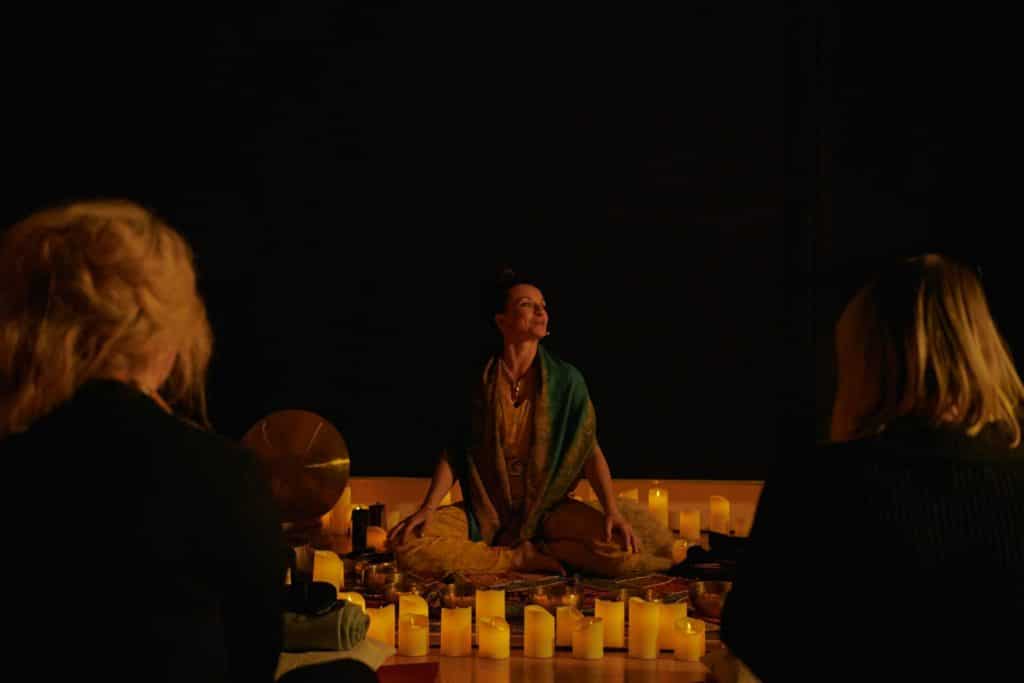 A woman in the center of the image instructing a Mindful Glow class surrounded by candles and two guests watching her.