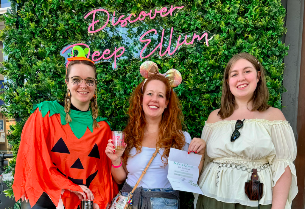 Image shows three people in costumes in front of a sign that says "Discover Deep Ellum"