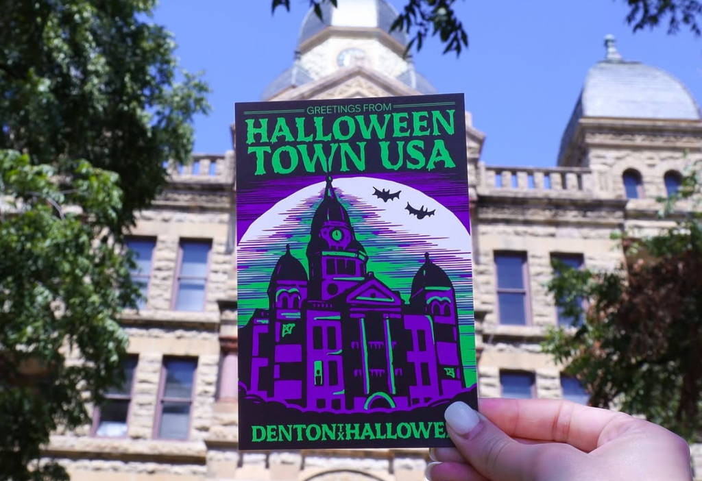 Image shows someone holding a sticker with the words "Halloween Town USA" in front of a Denton capital building.