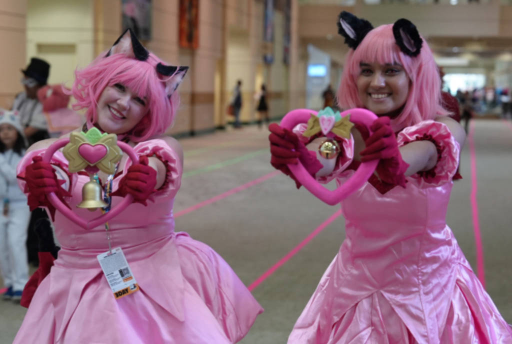 Image shows two cosplayers in costumes holding up a craft in the shape of a heart.