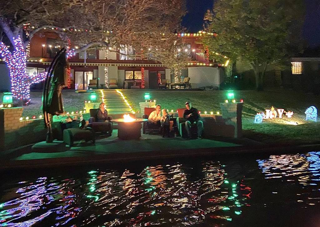 Image shows a group of people hanging out at the edge of a lake with a holiday-decorated home in the background.