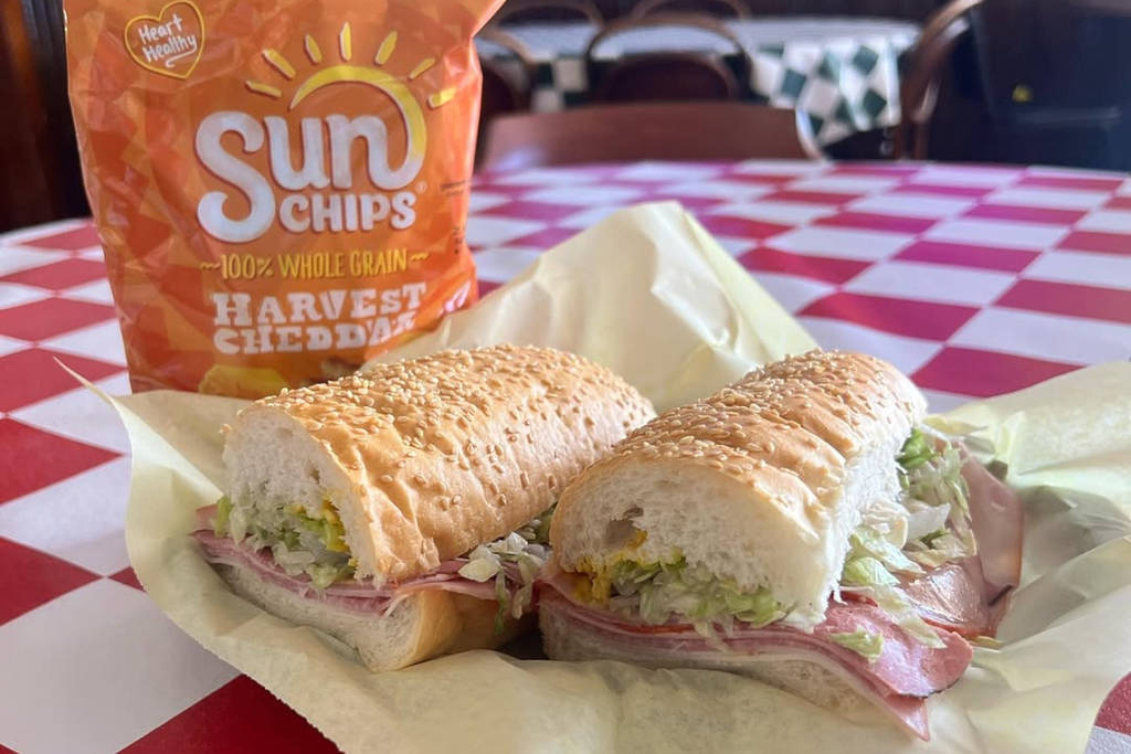 Image shows a sandiwch and bag of Sun Chips place on top of a checkered tablecloth inside DIno's Subs.