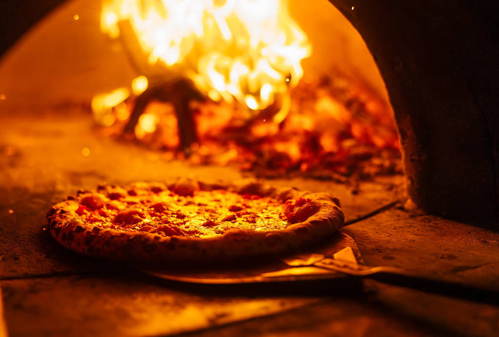 Image shows a pizza coming out of a wood-fired oven