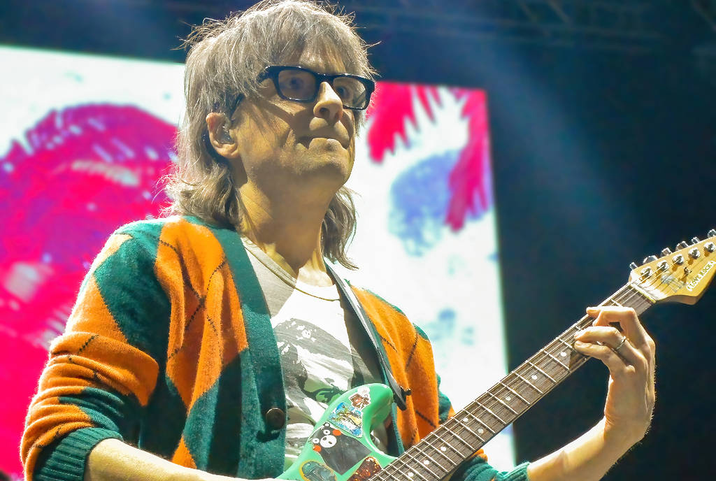 Image shows River Cuomo performing on stage in a cardigan.