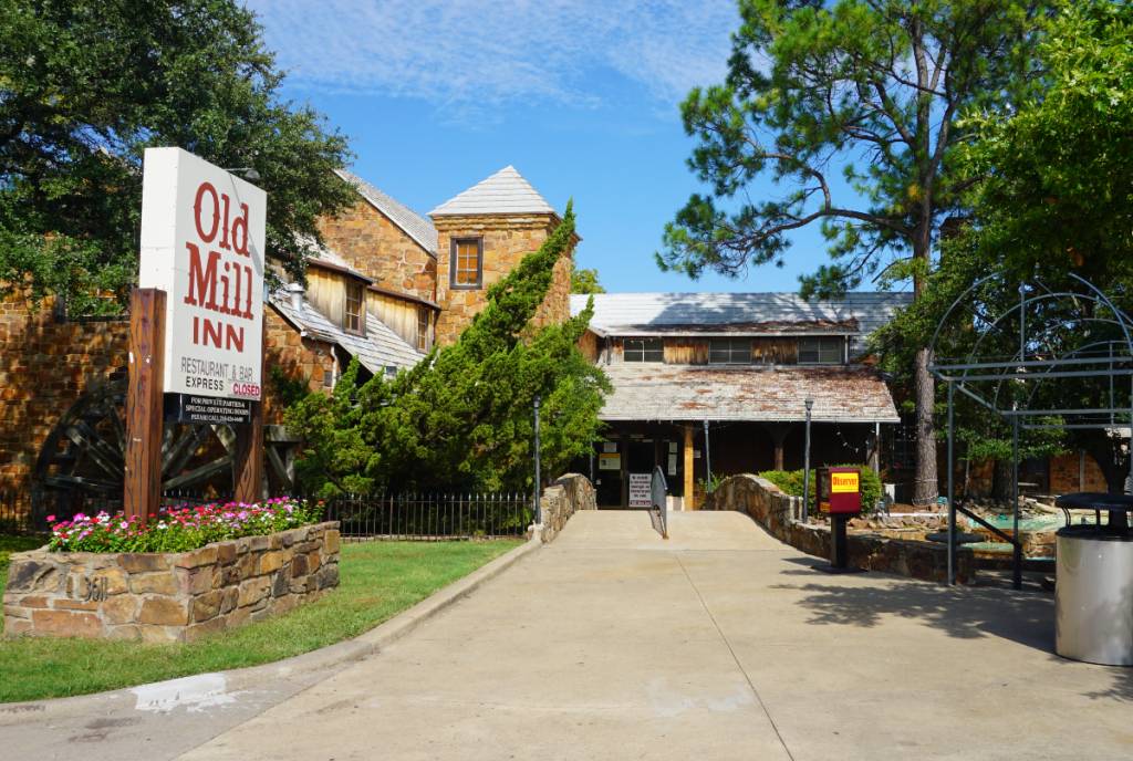 La imagen muestra el Old Mill Inn en Fair Park, Dallas, incluyendo el letrero y la fachada principal.