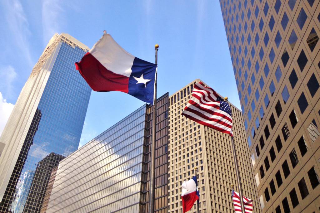 La imagen muestra las banderas de Texas y de EE. UU. ondeando con edificios al fondo.