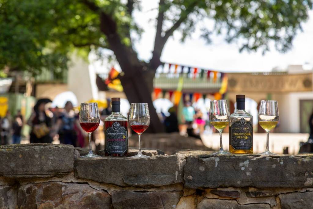 Image shows different glasses of mead with a festival atmosphere in the background. 