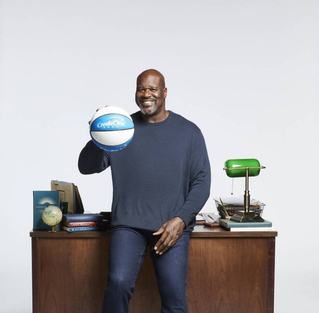 Image shows Shaquille O'Neal sitting on a desk for a posed photograph.