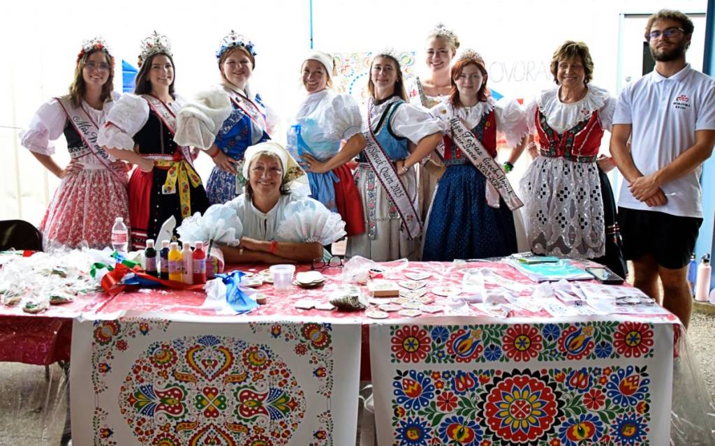 La imagen muestra a un grupo de personas vestidas con trajes tradicionales checos reunidas alrededor de una mesa. 