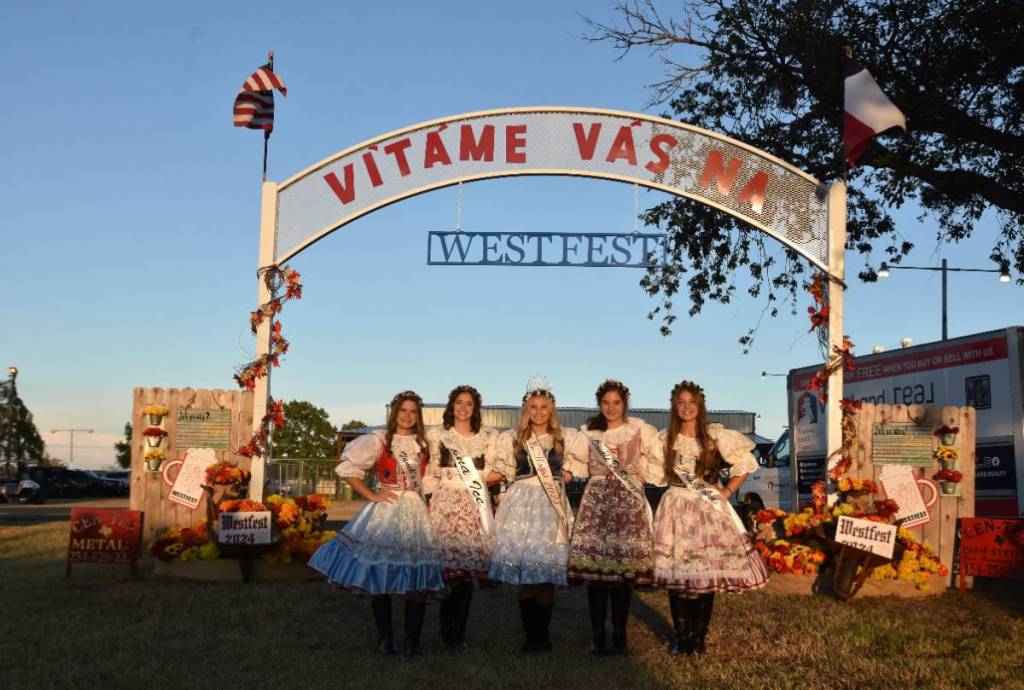 La imagen muestra a un grupo de chicas con trajes tradicionales checos posando debajo de un cartel escrito en checo. 