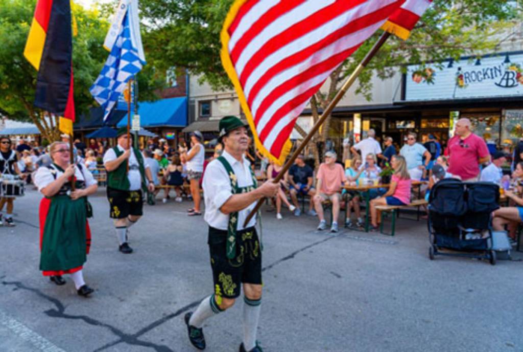 Image shows a man dressed in traditional German attire holding up an American flag.