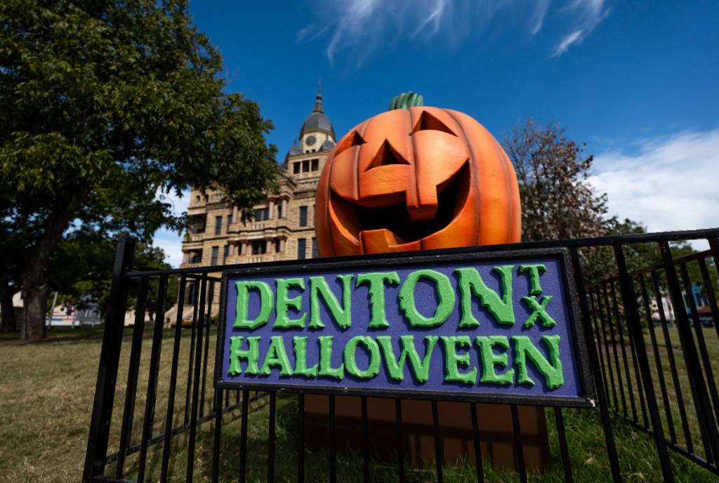 Image shows a jack-o-lantern installation propped up over a sign that reads "Denton Halloween" with the Denton courthouse in the background.