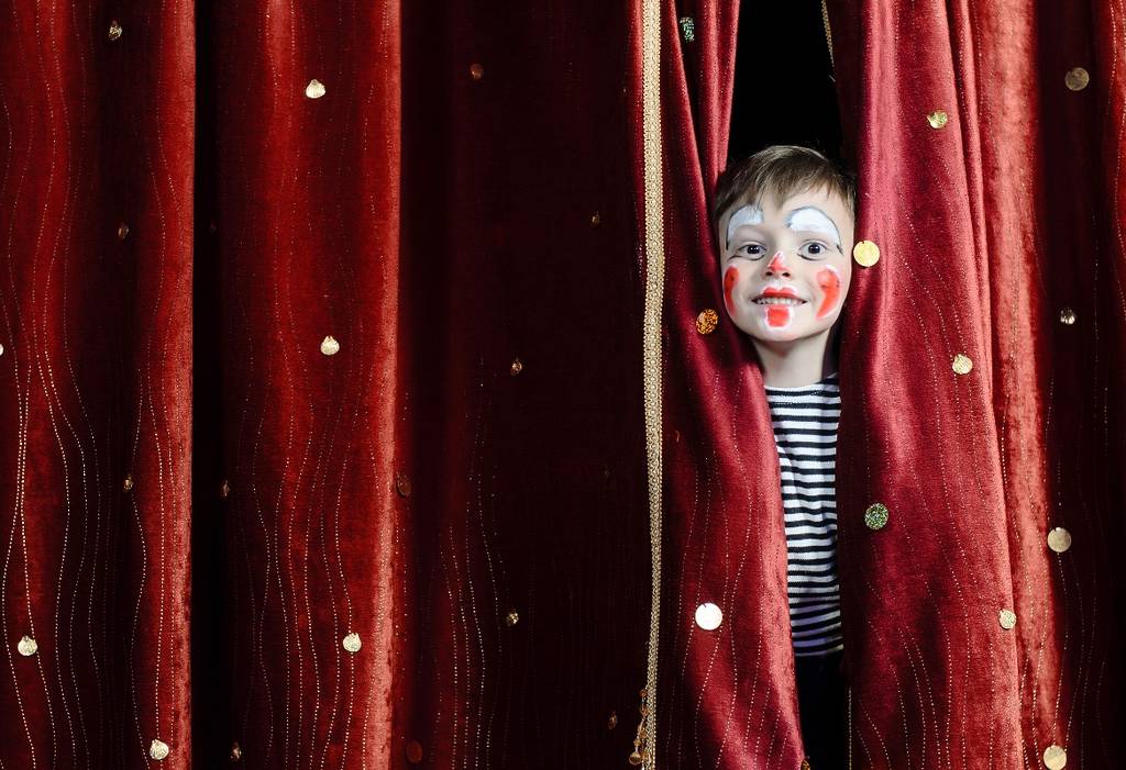 Little boy behind the red curtain in the theater wearing clown makeup