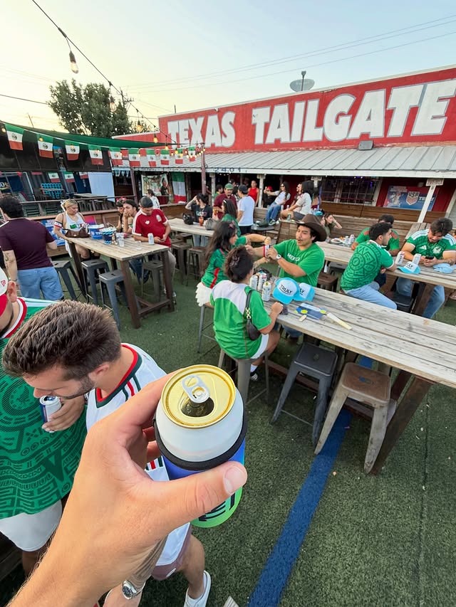La imagen muestra a una persona levantando una cerveza en medio de un grupo de gente en el Texas Tailgate durante una fiesta para ver el partido de México. 