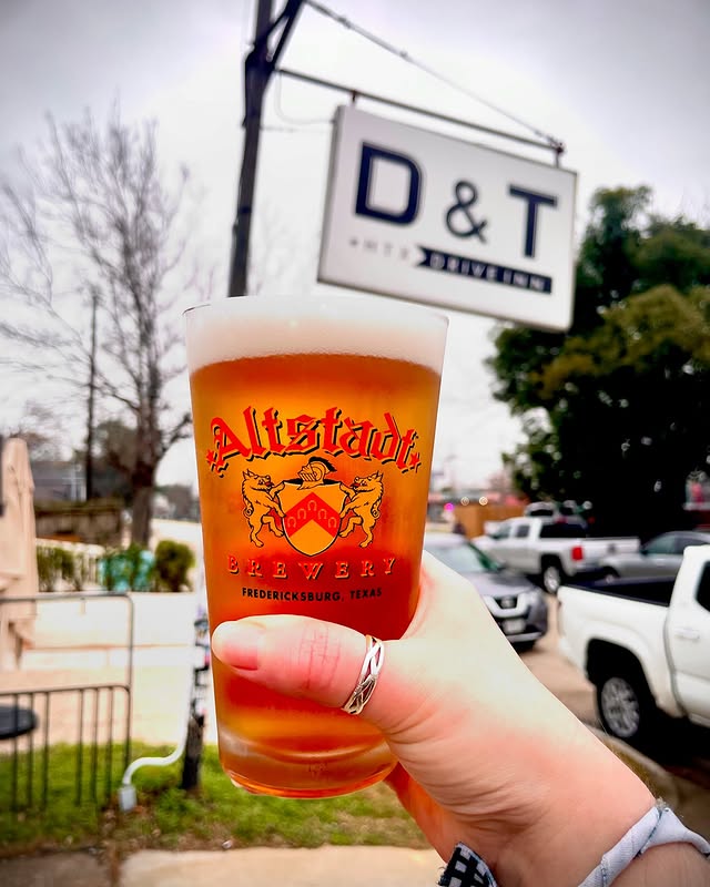 La imagen muestra a una persona sosteniendo un vaso de cerveza con el letrero del D&T Drive Inn al fondo. 