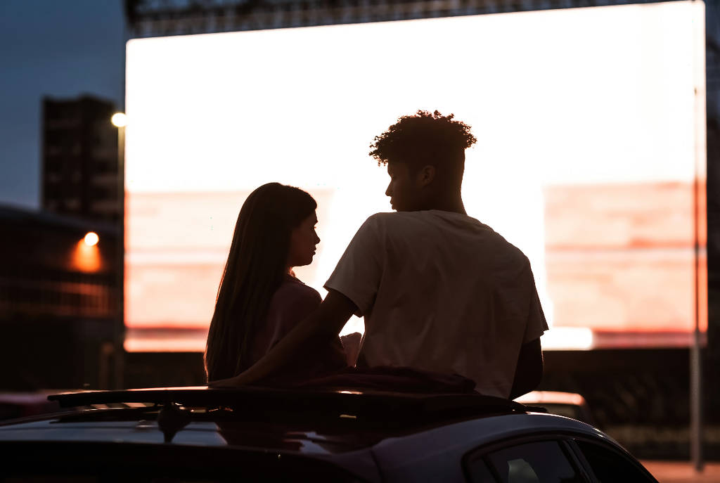 Image shows a young couple in front of a screen at a drive-in theater.