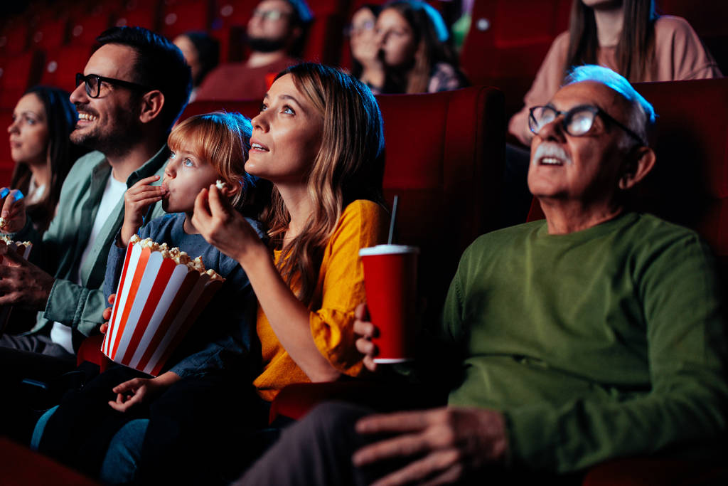 Image shows a group of theater-goers inside a theater watching the screen with popcorn and drinks.