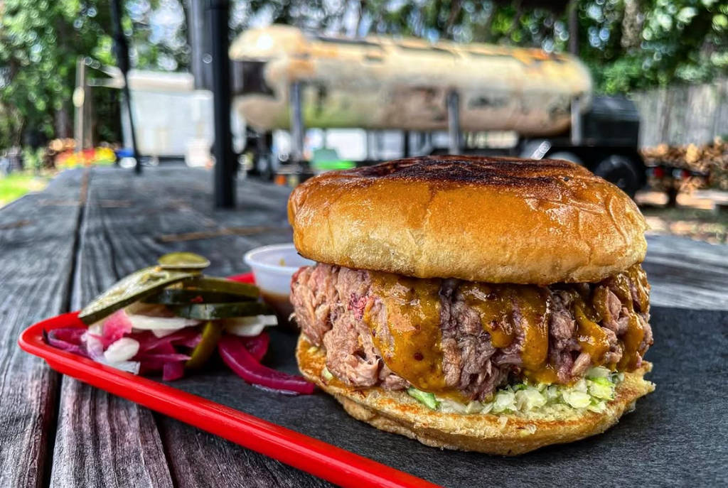 Image shows a barbecue sandwich with a smoker in the background at Rosemeyer Bar-B-Q in Houston, Texas