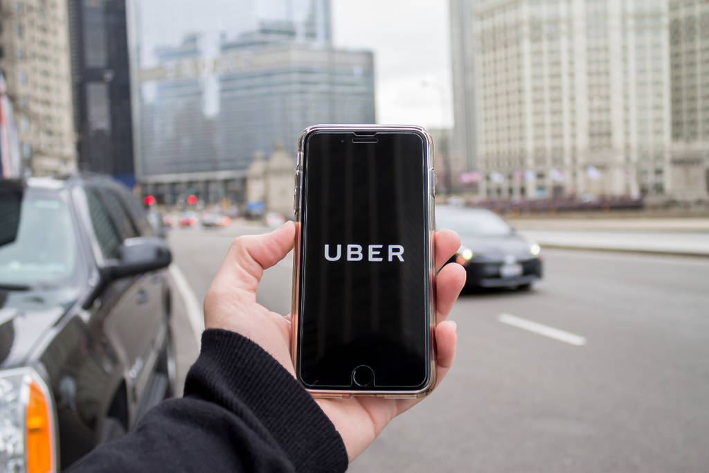 Image shows someone holding up their phone with the Uber app featured on the screen with a cityscape in the background.