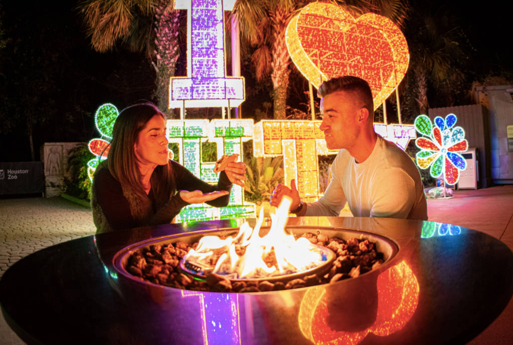 La imagen muestra a una pareja comiendo s'mores alrededor de una hoguera con una instalación de luces de fondo en la experiencia Zoo Lights, sólo para adultos, del zoo de Houston.