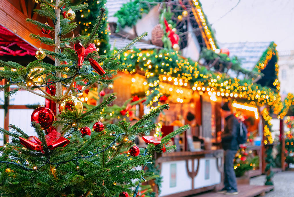 Image shows a Christmas tree with a decorated vendor in the background at a Christmas market.