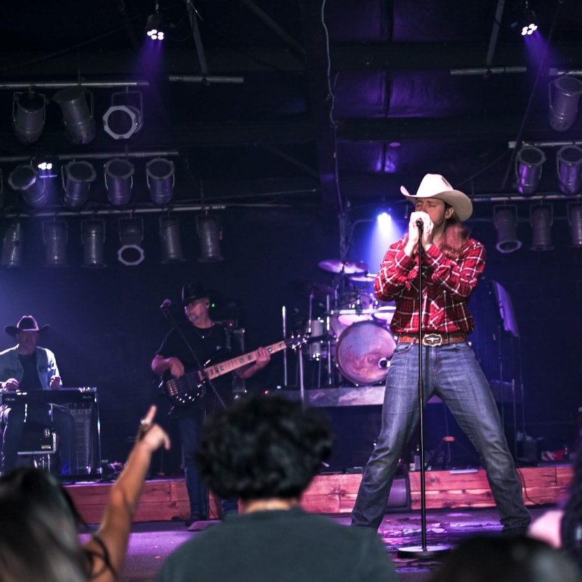 Image shows an artist in country ware singing during a live performance on stage at Cowboy Red River in Dallas.