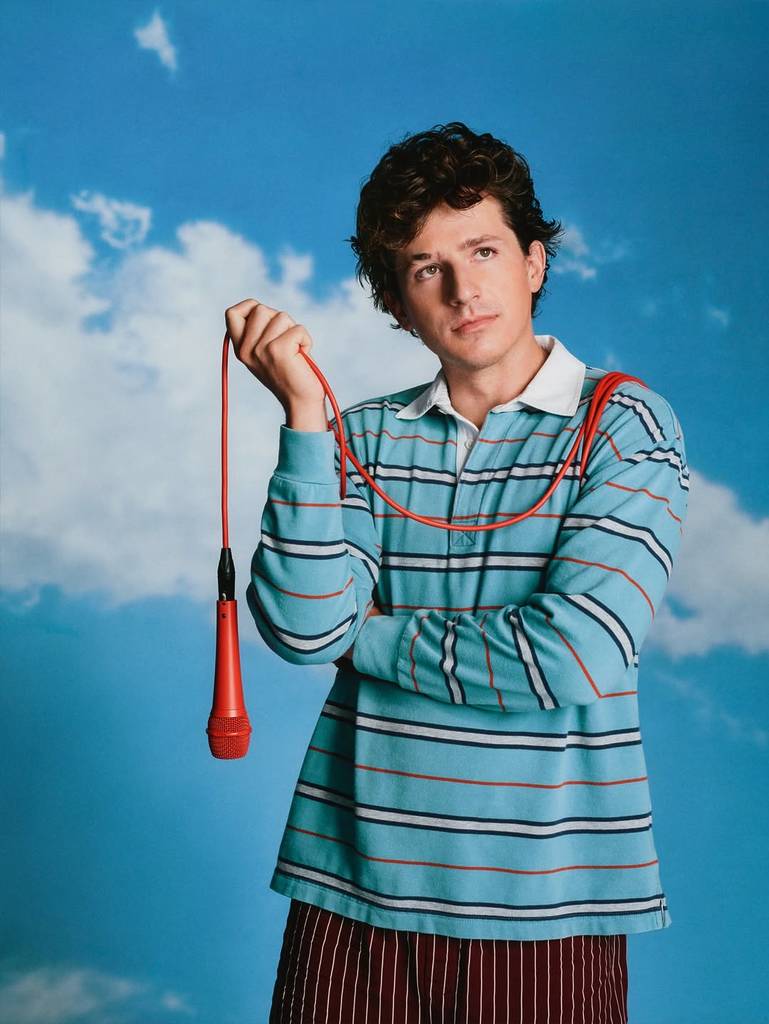 Image shows Charlie Puth posing with a microphone slung over his shoulder against a sky backdrop.