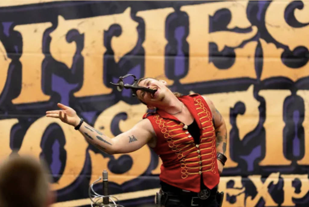 Image shows a performer singing into a microphone in front of a banner for the Oddities & Curiosities Expo
