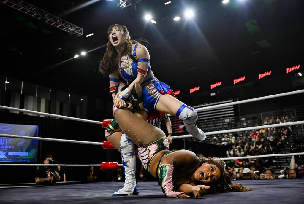 Image shows two TJPW wrestlers during a bout with one in a blue dress costume holding the other in a sort of leg-lock maneuver.