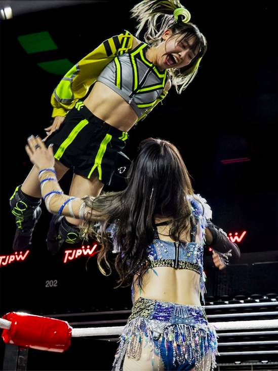 Image shows TJPW wrestlers during a bout with one leaping on to the other in a flying maneuver.