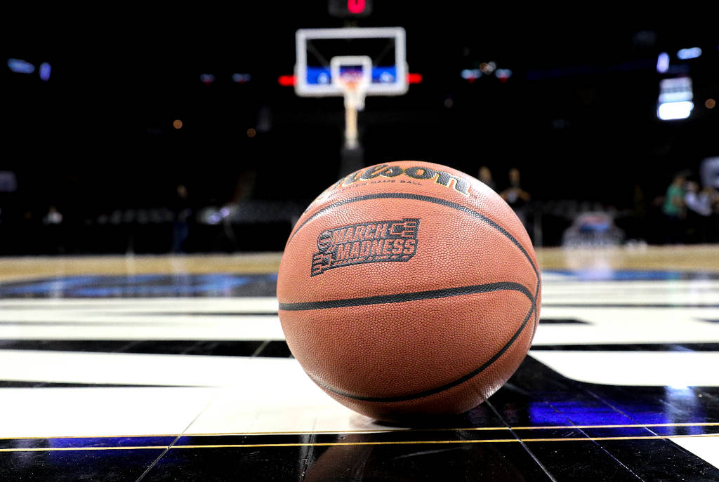 La imagen muestra una pelota de baloncesto de March Madness en la cancha con una canasta al fondo.
