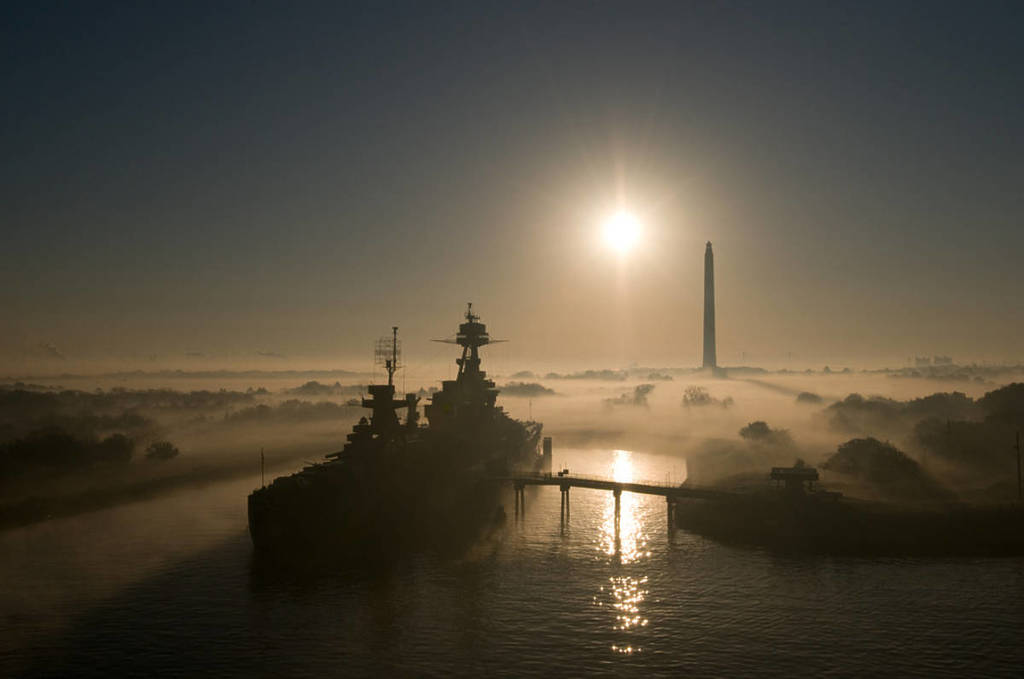 Image shows the USS Texas battleship in port with the San Jacinto Monument in the background in a foggy atmosphere. 