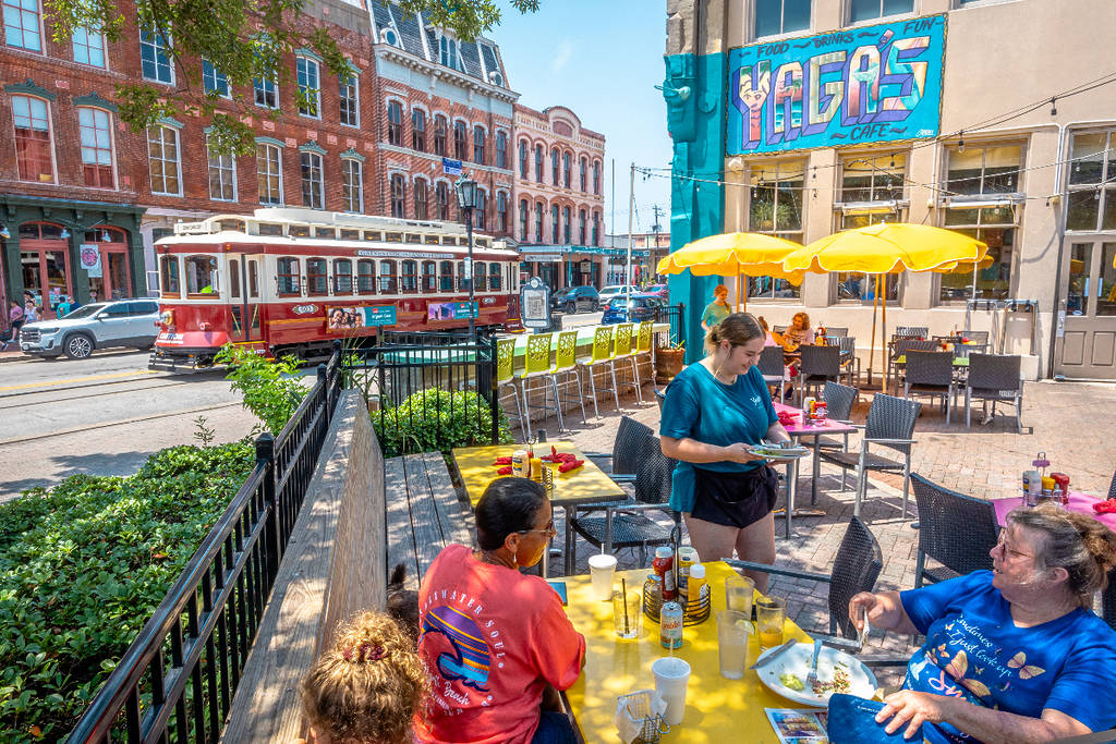 Image shows an outdoor patio in Galveston populated by patrons with a trolley in the background in Galveston. 
