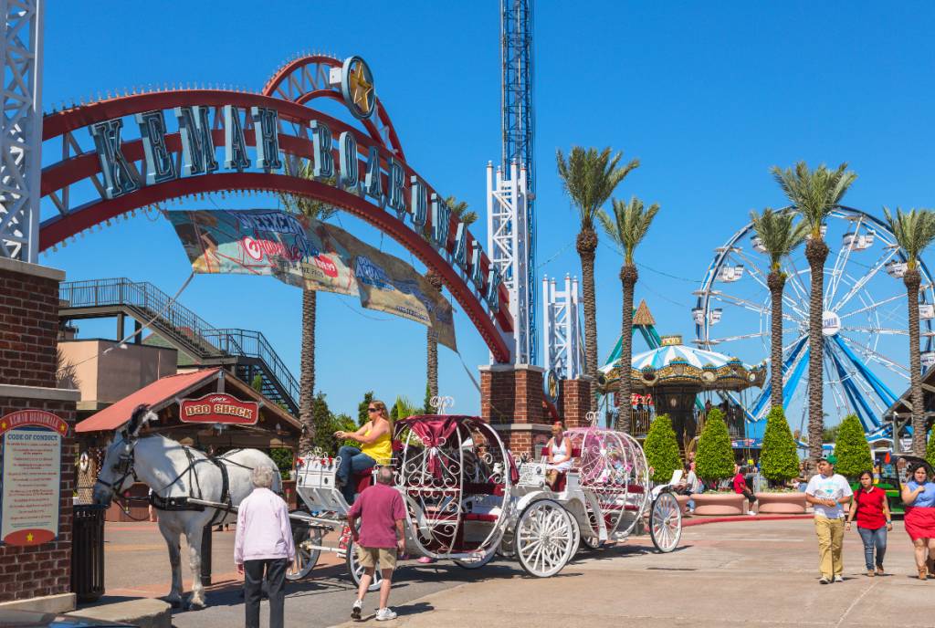 La imagen muestra la entrada principal del Kemah Boardwalk, con un carruaje, una noria y peatones.