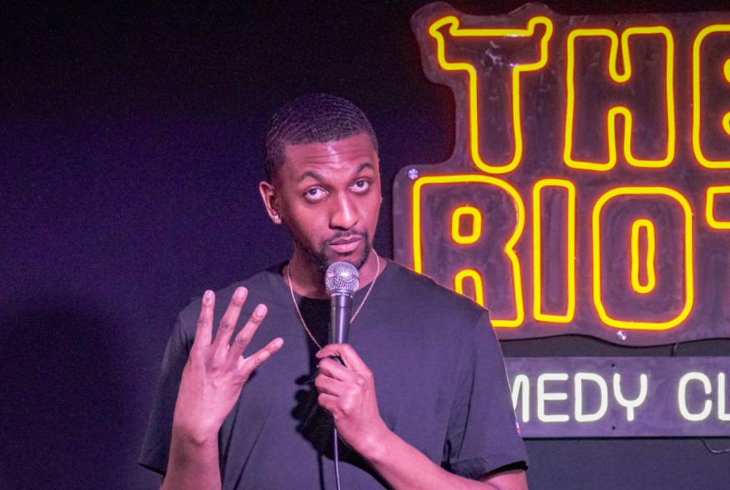 Image shows a comedian performing stand-up in front of "The Riot Comedy Club" signage. 
