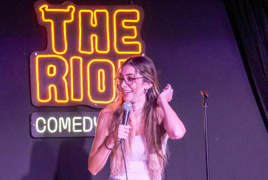 Image shows a female comedian stand-up performing in front of a neon sign that says "The Riot". 