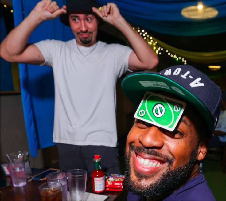 Image shows two men at a UNO Social Club one with a UNO card on his face the other in the background fixing his hat. 