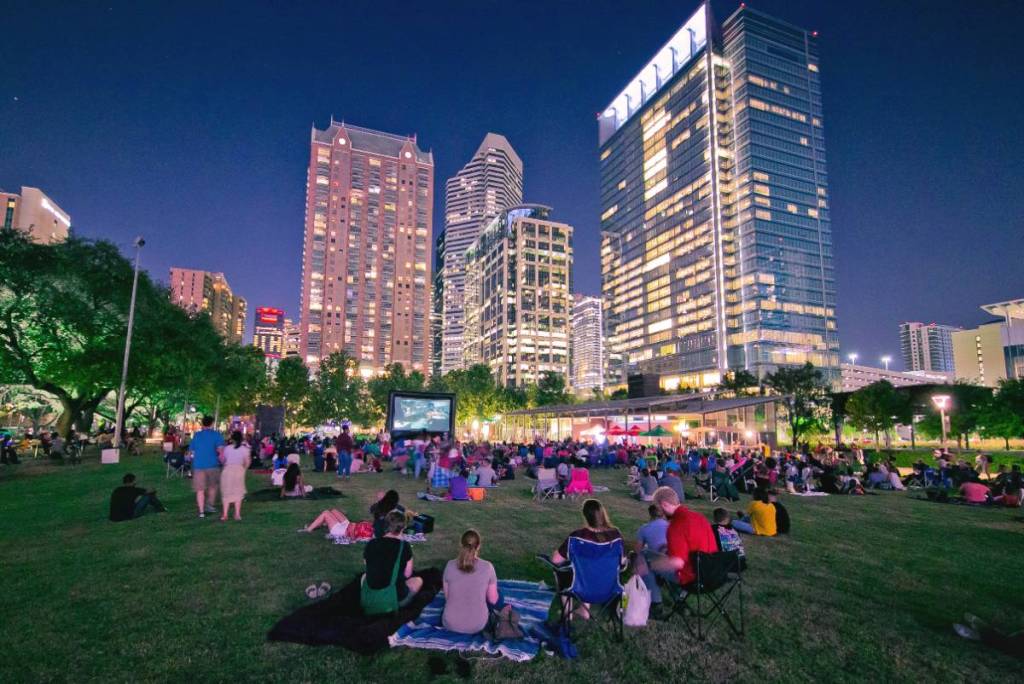 La imagen muestra a un grupo de personas viendo películas al aire libre en Houston, en el «Screen on the Green» de Discovery Green.