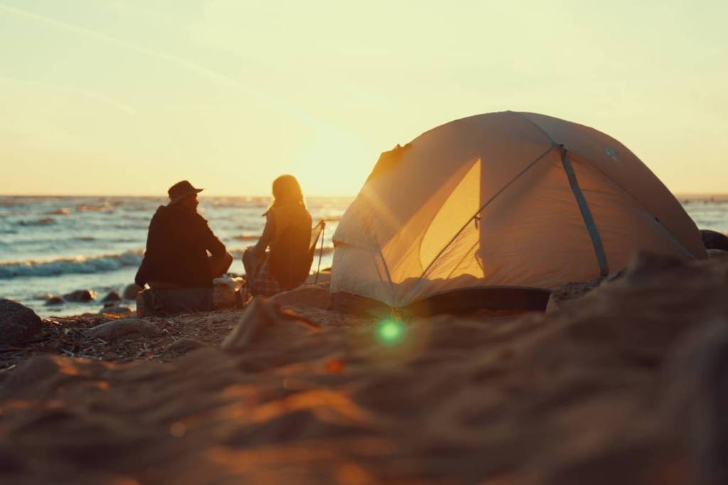 La imagen muestra a una pareja acampando en la playa junto a una tienda de campaña. 