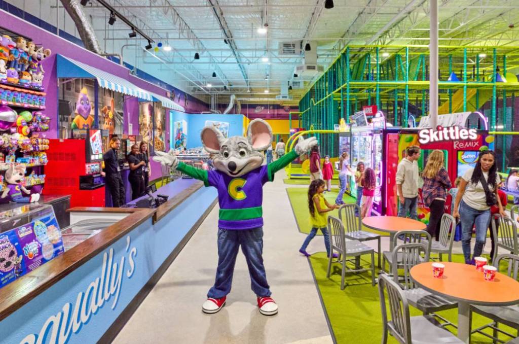 La imagen muestra a un personaje disfrazado de Chuck E. Cheese dentro de un Chuck E. Cheese