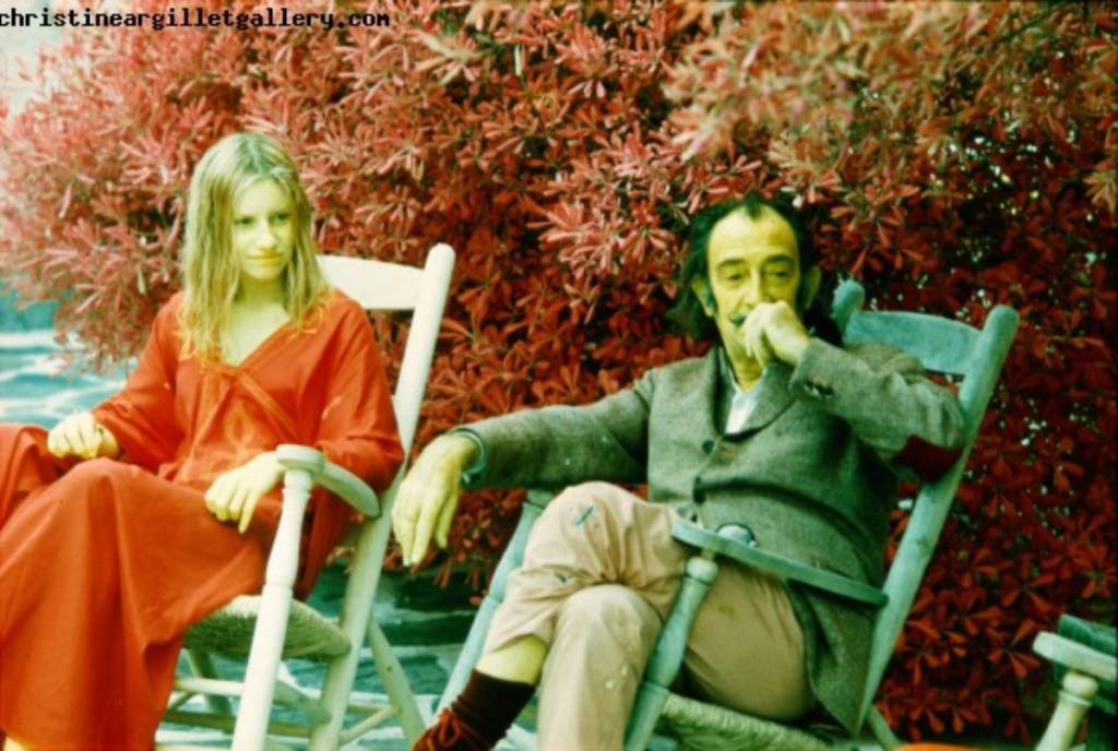 La imagen muestra a Salvador Dalí y a Christine Argillet sentados en unas tumbonas frente a un arreglo floral.