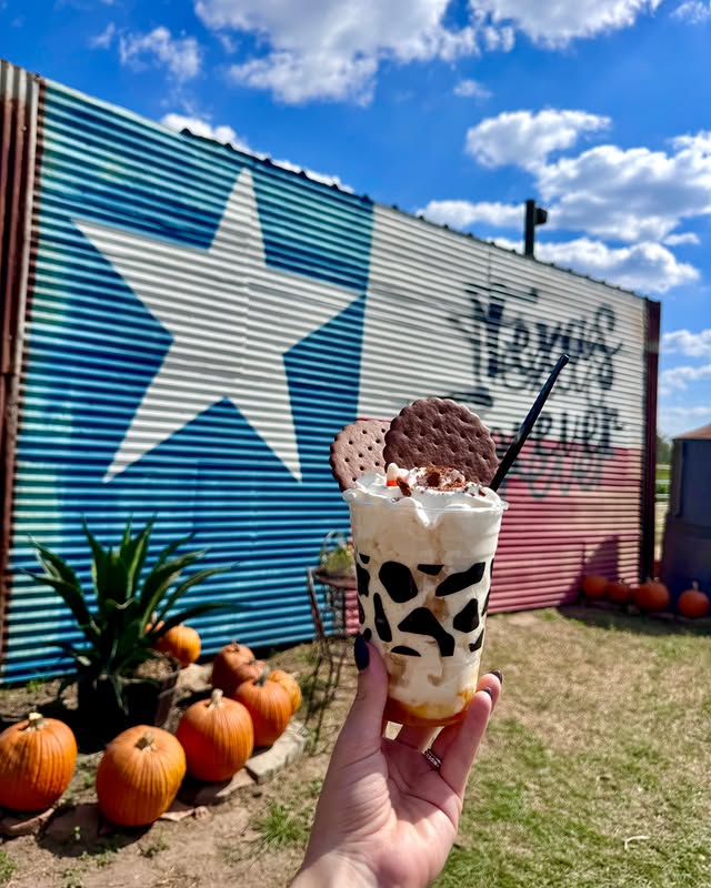 La imagen muestra a alguien fuera de plano sosteniendo una copa de postre con un edificio pintado con el diseño de la bandera de Texas y calabazas al fondo.