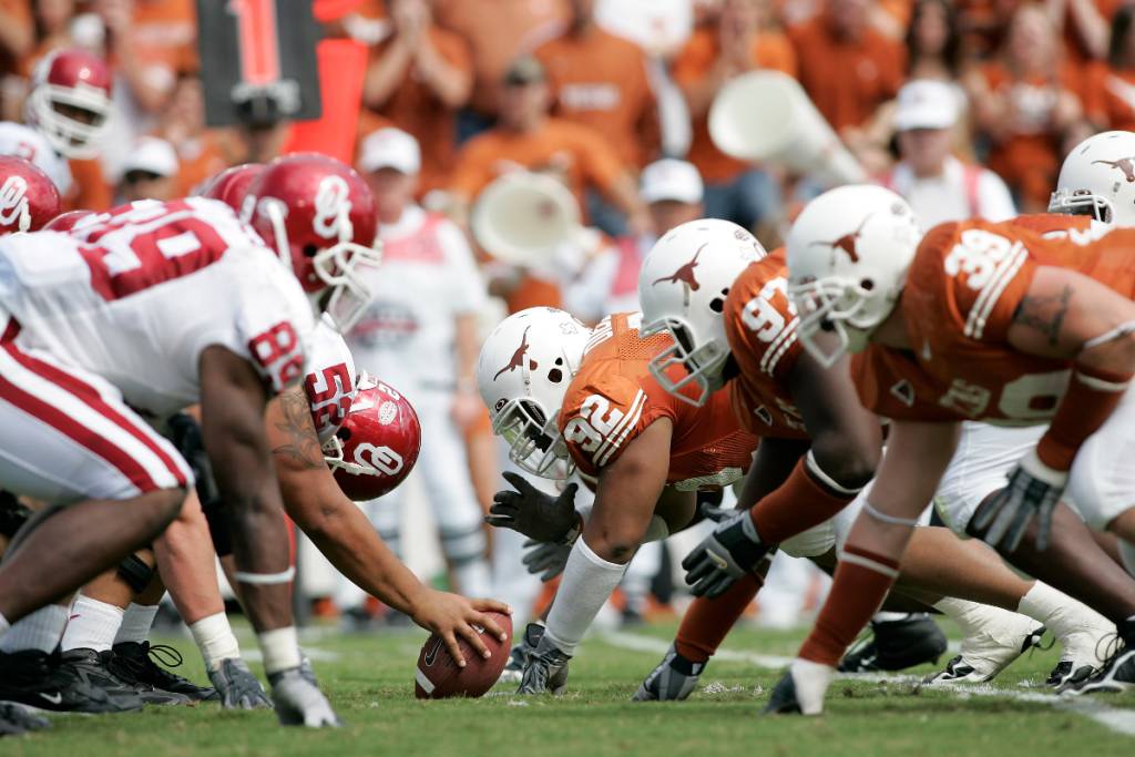 La imagen muestra la «Red River Rivalry» con los jugadores de fútbol americano de Oklahoma y de la Universidad de Texas alineados uno frente al otro.